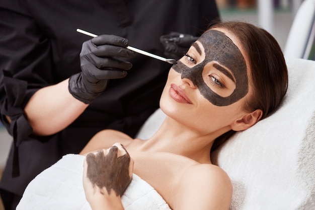 Cosmetologo che applica maschera nera sul viso di donna che indossa guanti neri, splendida donna nella spa con procedure facciali