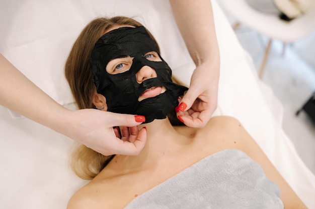 Cosmetologo che applica una maschera idratante nera sul viso della donna nella procedura di bellezza del salone di bellezza per