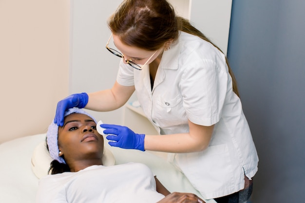 Cosmetologo donna al lavoro. Pelle di pulizia dell'estetista sulle palpebre attenta. Giovane donna africana che si trova sullo strato nella sala. Cosmetologia e cura della pelle