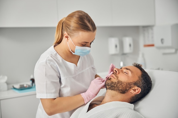 Cosmetologo medico femminile in maschera medica che fa iniezione nel viso maschile