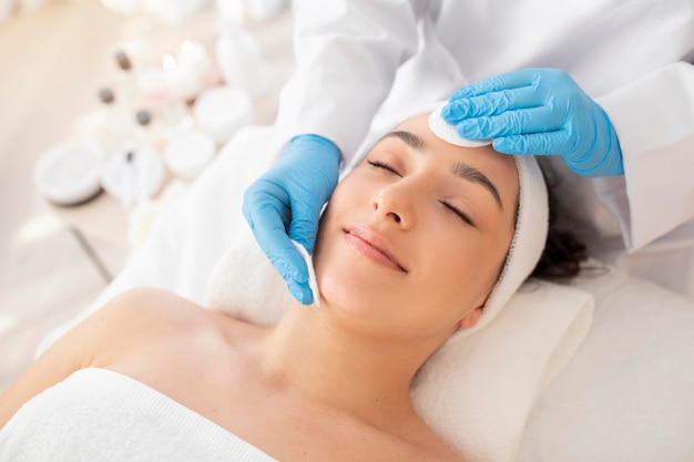 Cosmetologo per la cura del viso che utilizza tamponi di cotone per la pulizia della pelle della giovane donna indiana