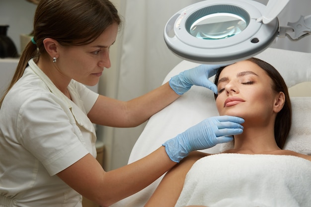 Cosmetologo professionista che lavora con il viso di donna bella in guanti blu presso la clinica di dermatologia