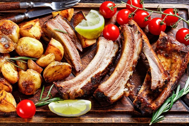 Costine di maiale alla griglia servite con patate novelle arrosto e pomodori freschi su un tagliere di legno su un tavolo di legno scuro vista orizzontale dall'alto