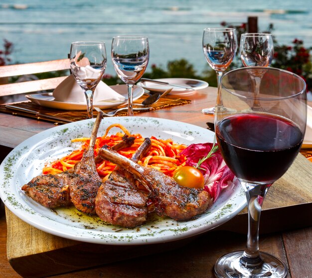 Costoletta di agnello con pasta e bicchiere di vino rosso con il mare in sottofondo