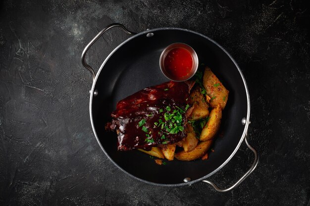 Costolette di manzo affumicate con patate arrosto rustiche, con salsa barbecue e condimenti, cipolle verdi in padella su un tavolo di pietra scura