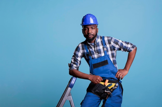 Costruttore serio appoggiato sulla scala che indossa tute e casco protettivo guardando la fotocamera su sfondo azzurro. Studio girato con il concetto di industria delle costruzioni.