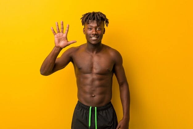 Costume da bagno da portare senza camicia del giovane uomo di colore che sorride mostrando numero cinque di mostra allegro con le dita.
