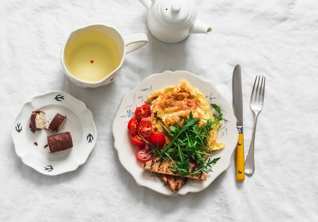 Cosy omelette da brunch fatta in casa con arugula ciliegia pomodori pane tostato integrale tè verde cioccolato dessert su uno sfondo chiaro vista dall'alto