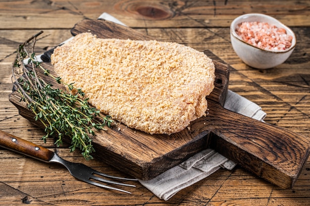 Cotoletta alla milanese impanata cruda tenera su una tavola di legno con timo. fondo in legno. Vista dall'alto.