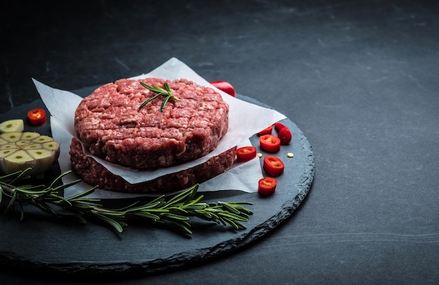 Cotoletta cruda dell'hamburger dalla carne di manzo con aglio e rosmarino su fondo nero con lo spazio della copia