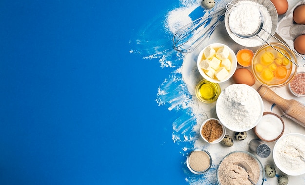 Cottura degli ingredienti per la pasta su sfondo di colore blu, vista dall'alto di farina, uova, burro, zucchero e utensili da cucina per la cottura in casa. Banner di concetto di cucina con copia spazio per il testo