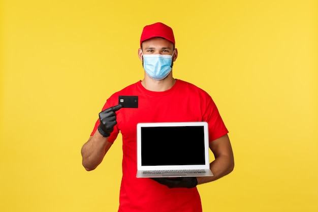 Covid-19, acquisto sicuro, consegna espressa e concetto di tracciamento degli ordini. Corriere sorridente in maschera medica, uniforme, mostrando carta di credito e pagina web sullo schermo del laptop, pagamento facile senza contatto