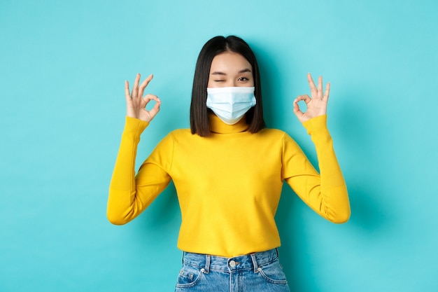 Covid-19, allontanamento sociale e concetto di pandemia. Allegra ragazza asiatica in mascherina medica che mostra gesti giusti, strizzando l'occhio alla telecamera, mostrando approvazione, in piedi su sfondo blu.