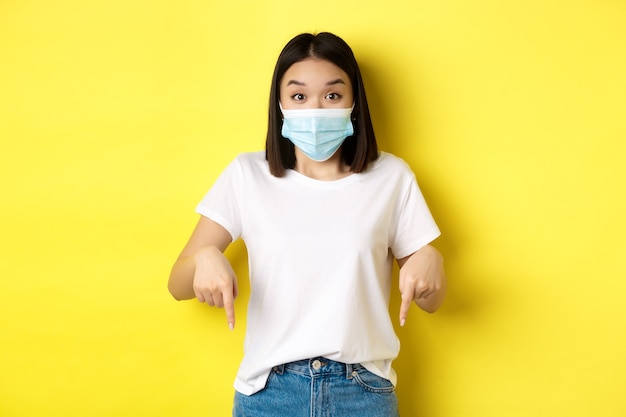 Covid-19, concetto di pandemia e allontanamento sociale. Donna asiatica stupita in mascherina medica, mostrando pubblicità, indicando a destra e sorridente, sfondo giallo.