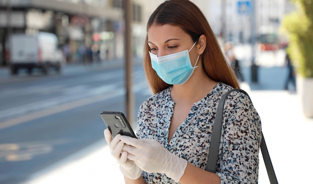 COVID-19 Donna che indossa una maschera chirurgica e guanti protettivi utilizzando l'applicazione per cellulare in una strada cittadina