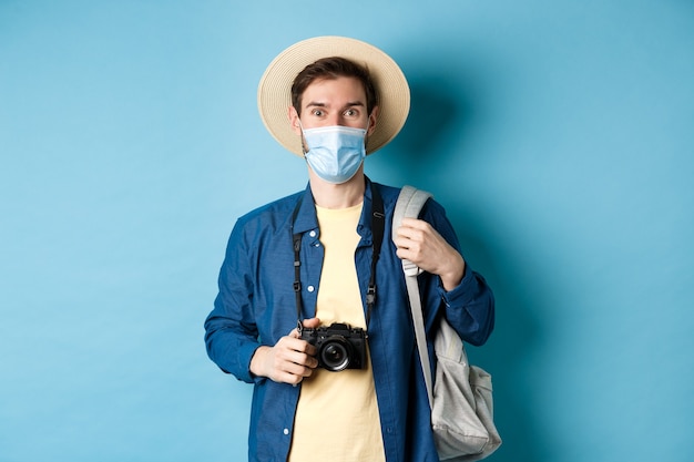 Covid-19 e il concetto di viaggio. Giovane turista in maschera medica e cappello estivo viaggia all'estero durante la pandemia di coronavirus, scatta foto in vacanza, sfondo blu.