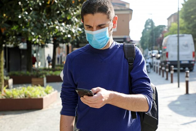 COVID-19 Giovane che indossa la maschera FFP2 utilizzando uno smartphone in una strada cittadina