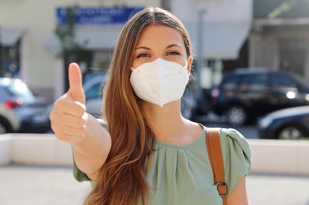 COVID-19 Giovane donna positiva che indossa la maschera protettiva FFP2 che evita il coronavirus 2019 che mostra i pollici in su in una strada cittadina