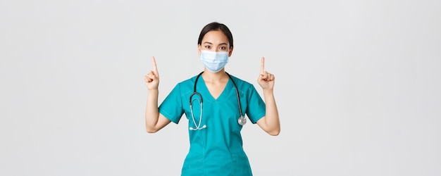 Covid-19, malattia da coronavirus, concetto di operatori sanitari. Giovane dottoressa asiatica professionista, infermiera con maschera medica e scrub, puntando il dito verso l'alto, mostrando la strada, pubblicità.
