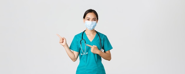 Covid-19, malattia da coronavirus, concetto di operatori sanitari. Medico femminile asiatico sicuro di sé, dottore in maschera medica e scrub che puntano le dita a sinistra, mostrando pubblicità.