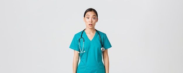 Covid-19, operatori sanitari, concetto di pandemia. L'infermiera asiatica impressionata e sorpresa in camice lascia cadere la mascella e fissa la telecamera senza parole, guardando stupita su sfondo bianco