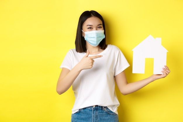 Covid-19, pandemia e concetto immobiliare. La donna asiatica allegra che sorride nella mascherina medica, mostrando il ritaglio della casa di carta, consiglia l'agenzia per l'acquisto della proprietà.