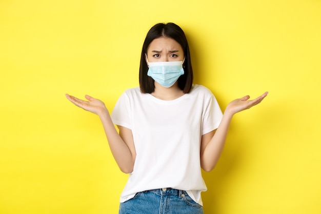 Covid-19, quarantena e concetto di distanza sociale. Donna asiatica confusa in maschera medica che alza le spalle, allarga le mani di lato all'oscuro, non sa nulla, sfondo giallo