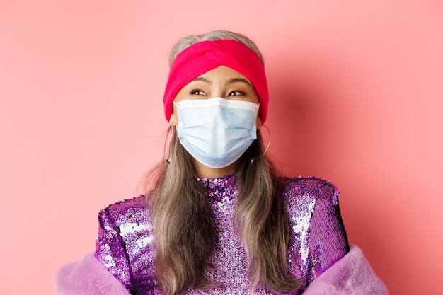 Covid-19, virus e concetto di moda. Primo piano di una donna anziana asiatica felice ed elegante in maschera medica che ride, guardando a sinistra allegra, in piedi su sfondo rosa.