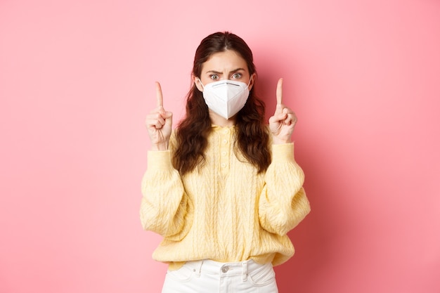 Covid, corona e concetto di allontanamento sociale. Giovane donna confusa e scioccata in respiratore medico che punta il dito verso l'alto, fissando spaventata la fotocamera, muro rosa