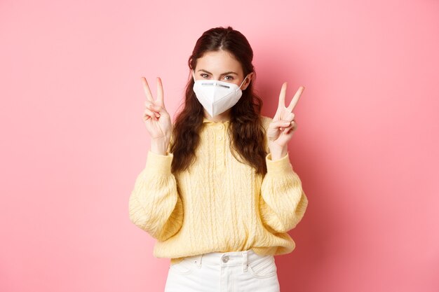 Covid lockdown e concetto di pandemia carina ragazza glamour indossa un respiratore medico per andare all'aperto durante la quarantena mostra pace vsign sta contro il muro rosa