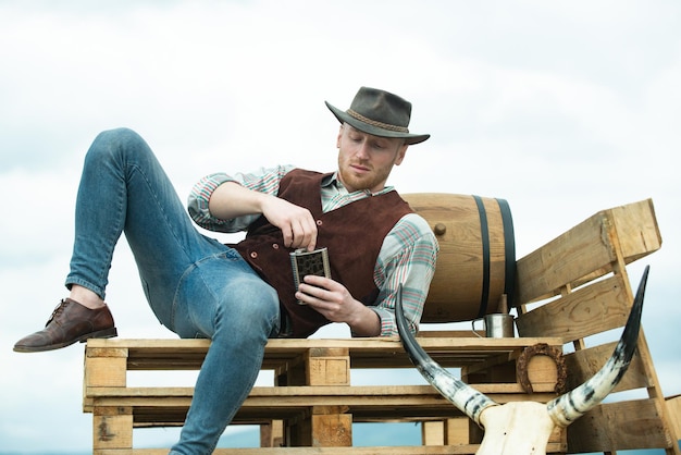 Cowboy agricoltore che indossa cappello vita occidentale paese americano ritratto maschile proprietario di una fattoria lavoratore in campagna in fattoria o ranch uomo attraente con whisky o brandy