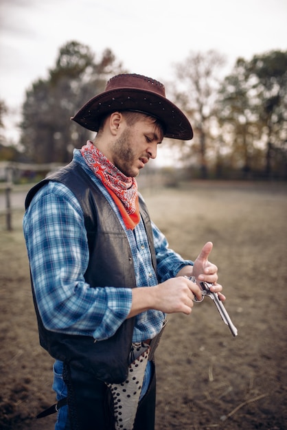 Cowboy controlla il suo revolver prima dello scontro a fuoco nel ranch, western. Persona di sesso maschile vintage con pistola in fattoria, avventura nel selvaggio west