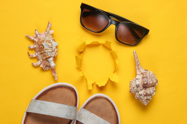 Creative summer beach flat lay. Sandali da donna in pelle, conchiglie, occhiali da sole gialli con foro strappato.