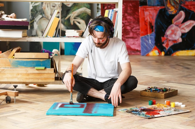 Creativo artista maschio hipster barbuto dai capelli scuri in cuffie e bandana blu seduto sul pavimento e disegno con olio e pennello su tela nel suo studio contemporaneo con molti dipinti