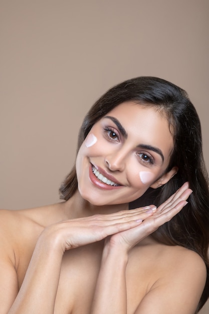 Crema delicata. Sorridente giovane donna dai capelli lunghi con crema sulle guance inclinando la testa e le palme a lato