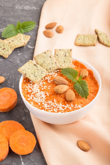 Crema di carote con semi di sesamo e snack in una ciotola bianca su uno sfondo nero di cemento. vista laterale, da vicino.