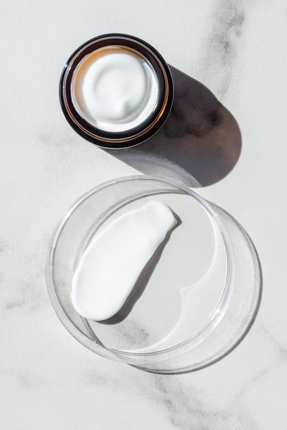 Crema per il viso in un barattolo e striscio di crema idratante nella capsula di Petri su sfondo di marmo bianco Bellezza del prodotto cosmetico per il viso vista dall'alto Lozione per la cura del corpo