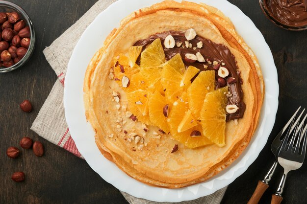 Crepes suzette con arance Crepes sottili con nocciole spalmabili al cioccolato e fette d'arancia frutta in piatto bianco per colazione su vecchio sfondo rustico tavolo in legno scuro Spazio di copia Vista dall'alto