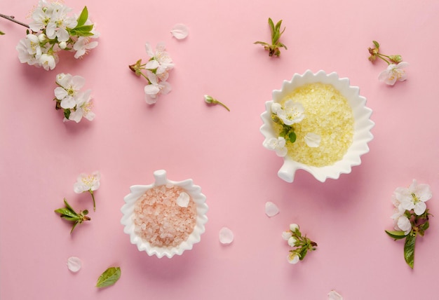 Cristalli di sale da bagno gialli in piatto di porcellana e fiori di ciliegio primaverili con foglie su sfondo di carta rosa per spa o cura personale