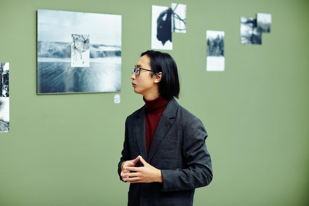 Critico d'arte professionista serio che indossa occhiali in piedi nella galleria d'arte moderna guardando foto in bianco e nero sul muro in mostra