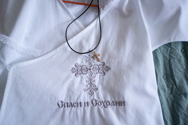 Croce ortodossa dorata su camicia da battesimo bianca per bambino con ricamo. accessori e gioielli religiosi tradizionali. l'ordinanza del battesimo. iscrizione in lingua russa - salva e conserva