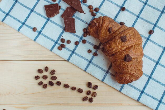 Croissant, chicchi di caffè e cioccolato su un tovagliolo da cucina e su uno sfondo di legno. La vista dall'alto.