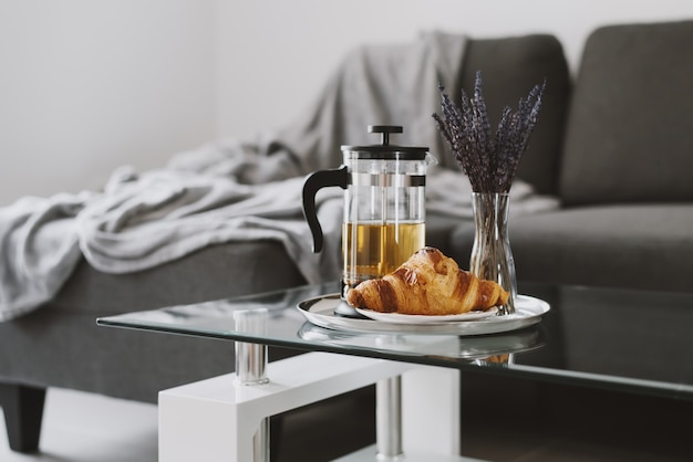 Croissant fresco e pentola per la stampa francese con tisana e vaso con lavanda su tavolino da caffè in vetro