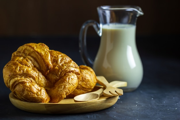 Croissant sul piatto e sulla brocca di latte di legno sulla tavola nel fondo scuro.