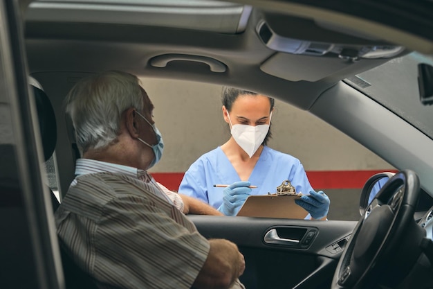 Crop medico femminile in maschera protettiva con clipboard contro il conducente maschile senior in automobile durante il check-up