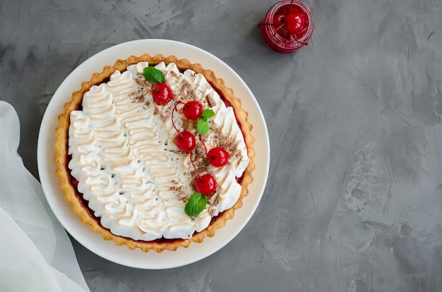 Crostata con ripieno di ciliegie e meringa italiana con ciliegina da cocktail, cioccolato e menta sopra su un piatto bianco su uno sfondo di cemento scuro. Torta di ciliegie. Orizzontale, copia dello spazio. Vista dall'alto.