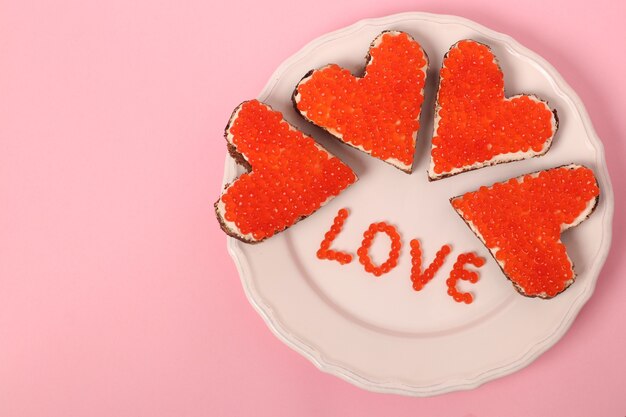 Crostino con caviale rosso e crema di formaggio a forma di cuore per San Valentino su sfondo rosa. Vista dall'alto