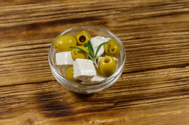 Cubetti di formaggio feta con olive verdi e rosmarino in ciotola di vetro su un tavolo di legno