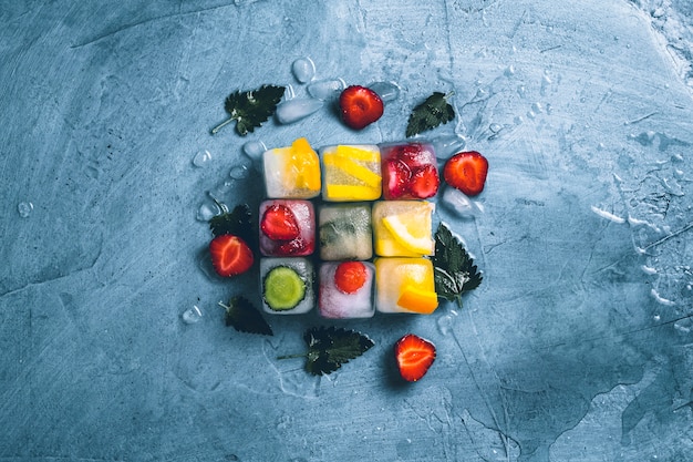 Cubetti di ghiaccio con frutta e ghiaccio rotto su uno sfondo blu di pietra con foglie di menta e frutta fresca. Forma del cubo. Menta, fragola, ciliegia, limone, arancia. Vista piana, vista dall'alto