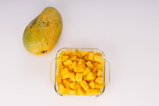 Cubetti di mango maturi o pezzi tagliati disposti in un contenitore quadrato di vetro con mango posizionato vicino su sfondo bianco.